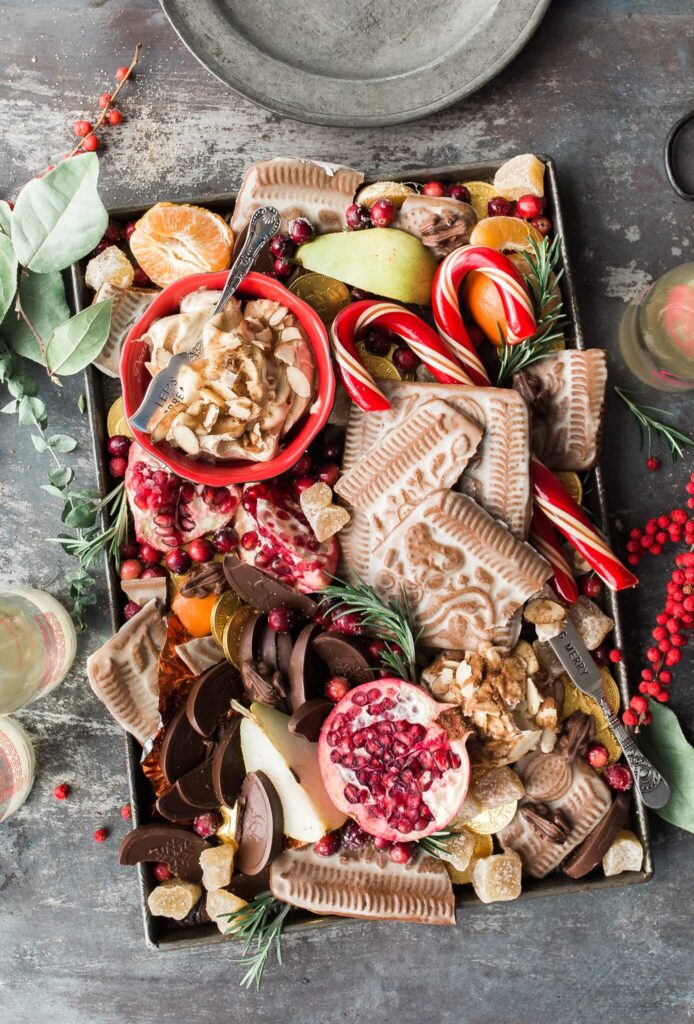 Against a grey background, a beautifully decorated tray of holiday goodies has candies, cookies, chocolates, and fruits on display.