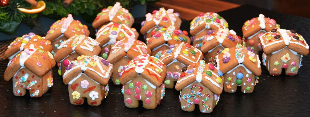Rows of mini, brightly decorated gingerbread houses sit in front of a holiday Christmas tree. The cookies are decorated with colorful candies and sprinkles with icing and frosting.