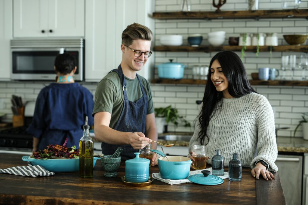 Three people are standing in a kitchen, cooking. One person has their back to the camera, two people face the camera. The people facing the camera are smiling and working with food on the counter.