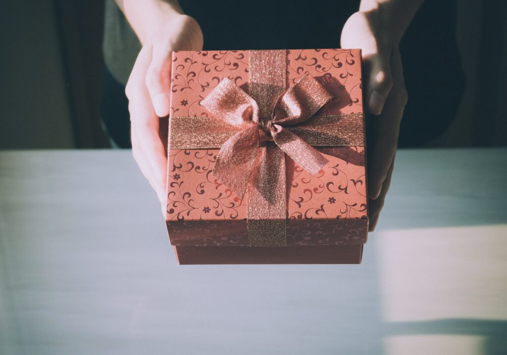 Two hands hold a pink or soft red gift box against a table background. The box is prettily decorated with metallic scroll patterns on the box itself as well as a shimmery ribbon tied around it.