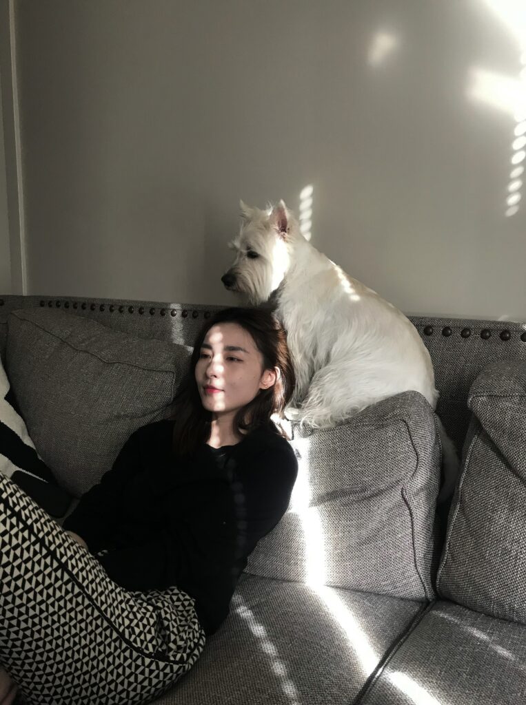 A woman is sitting on a couch watching a movie. Her dog is perched up on the back of the couch, half on her shoulder. 