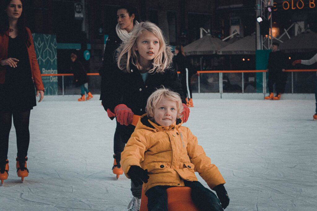 Two children are on an ice skating rink. The older sister is pushing her younger brother on a scooter.