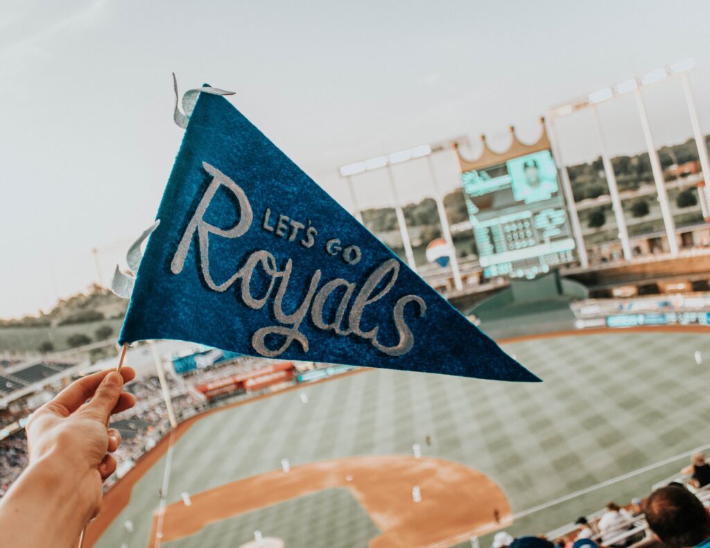 A hand is holding up a small, blue flag reading "Let's Go Royals" against Kauffman Stadium (located in Kansas City, Missouri) as the background. On select days, people can bring their dogs to watch the Royals play!