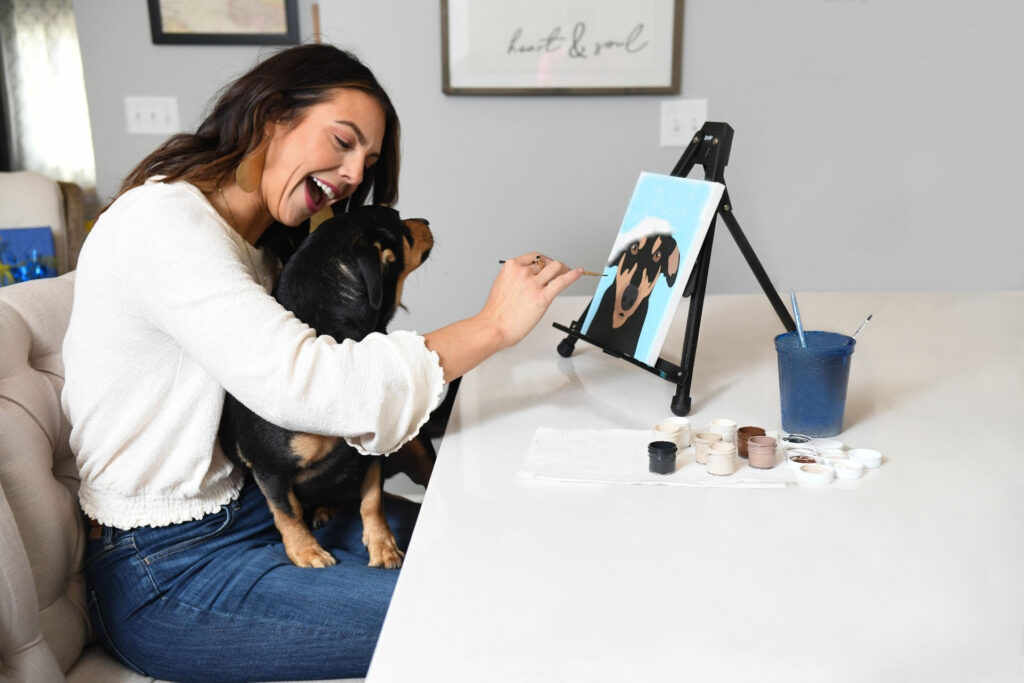 smiling happy woman with dog on lap while painting canvas of her dog